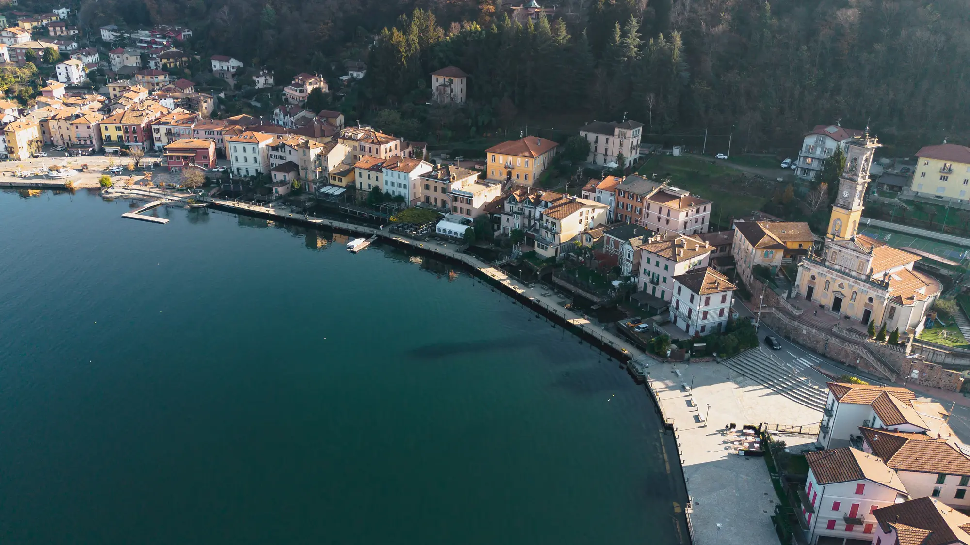 Aerial view of Porto Ceresio on Lake Lugano, showing the waterfront promenade, colorful houses, and surrounding hills